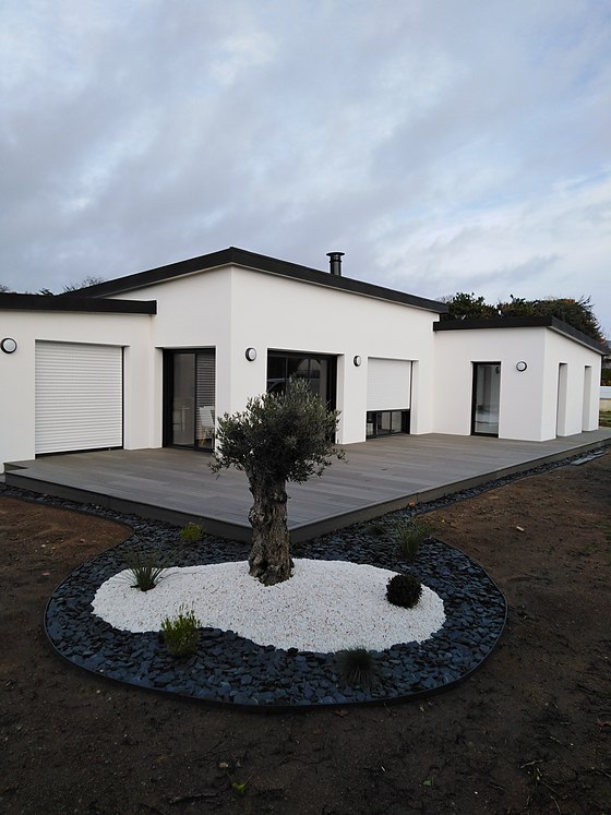 Terrasse composite grise périphérique autour d'une maison plain-pied contemporaine, jardin minéral avec olivier sur galets blancs.