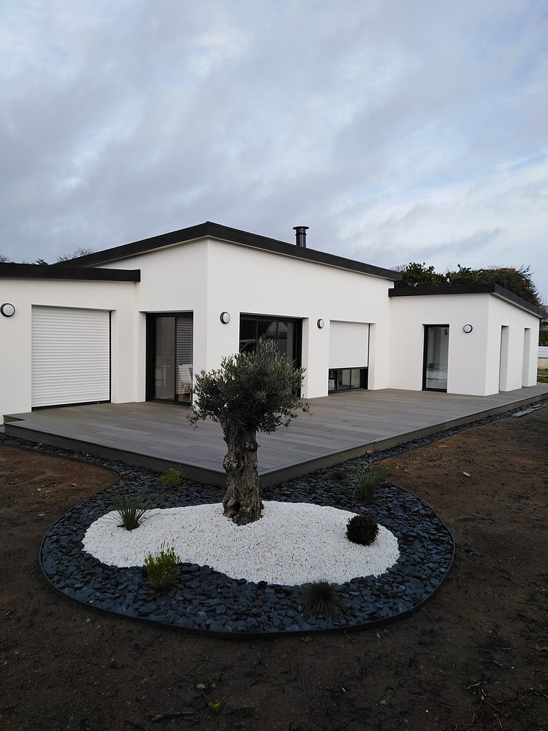 Terrasse composite grise périphérique autour d'une maison plain-pied contemporaine, jardin minéral avec olivier sur galets blancs.